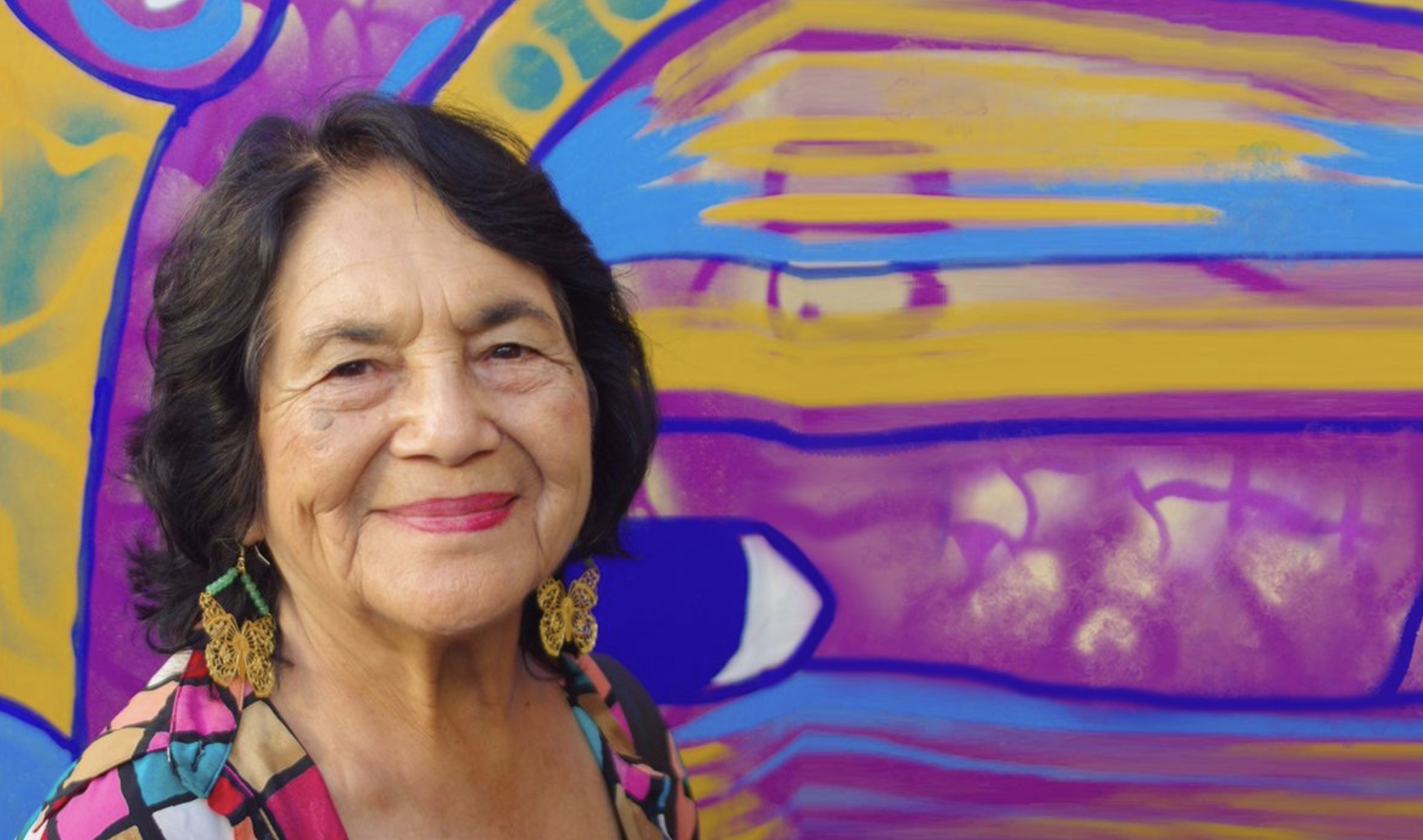 Dolores Huerta smiling in front of a vibrant mural