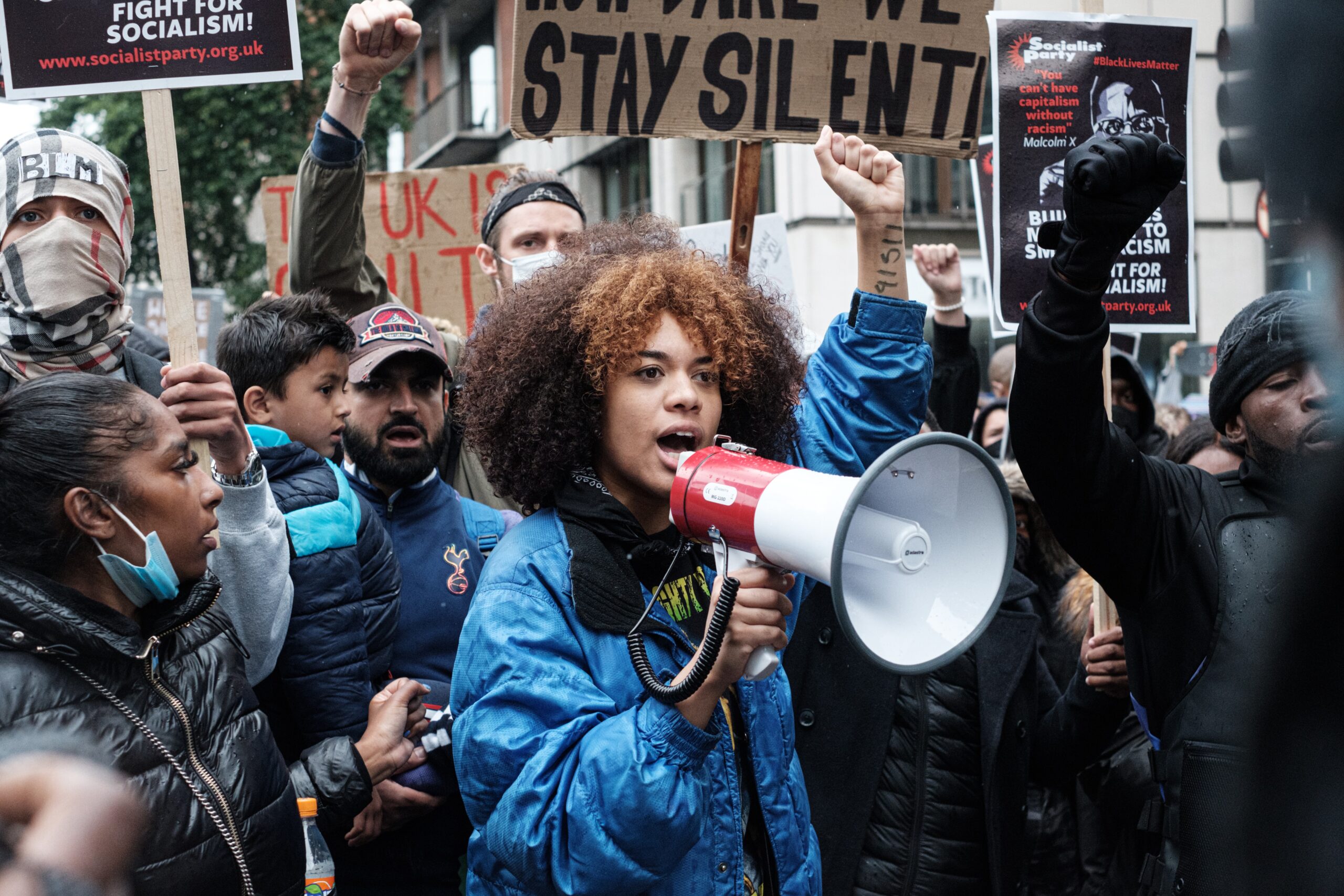 Behind a young woman with a megaphone, a crowd of protesters raise signs and their fists
