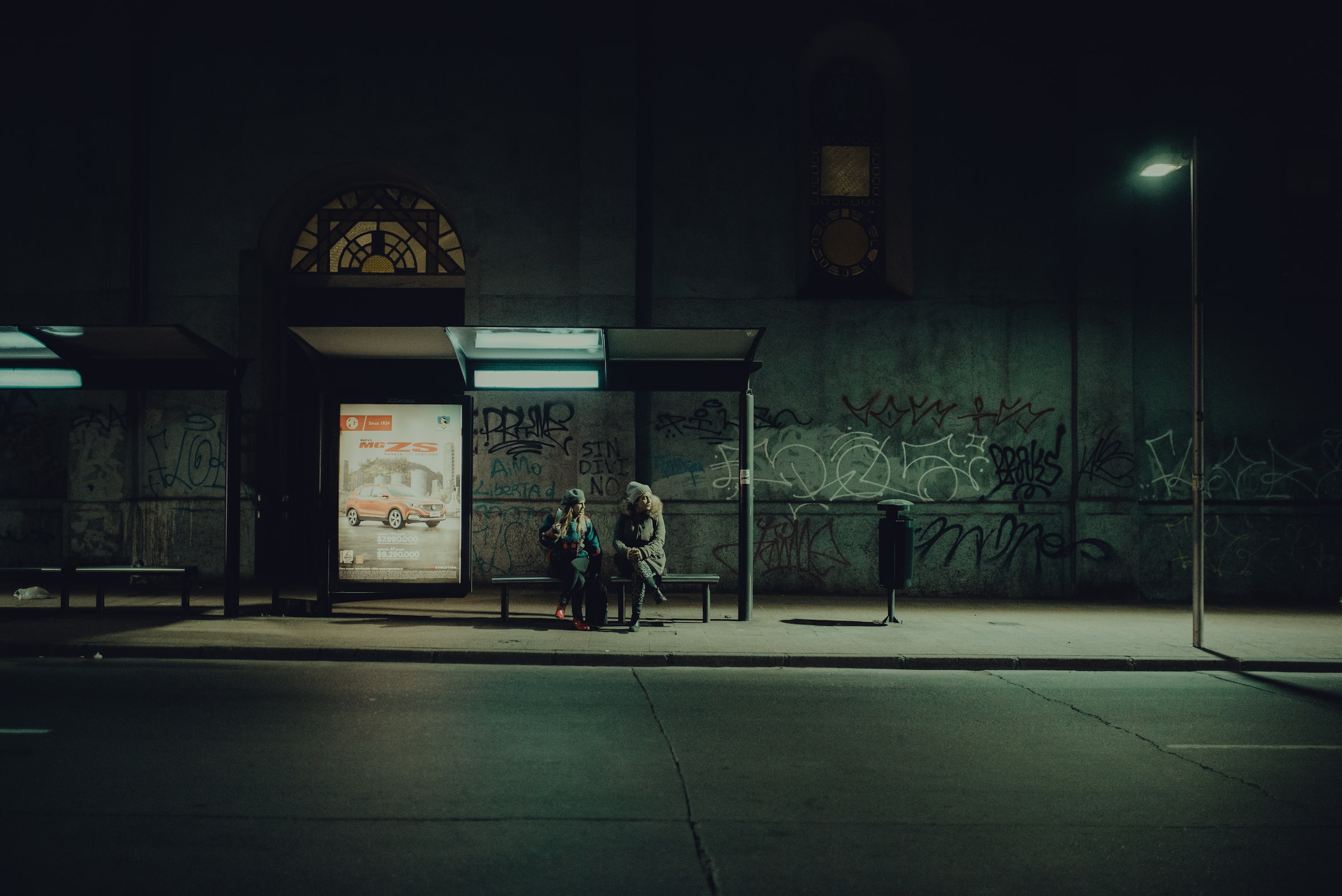 Two people sit at a bus stop at night