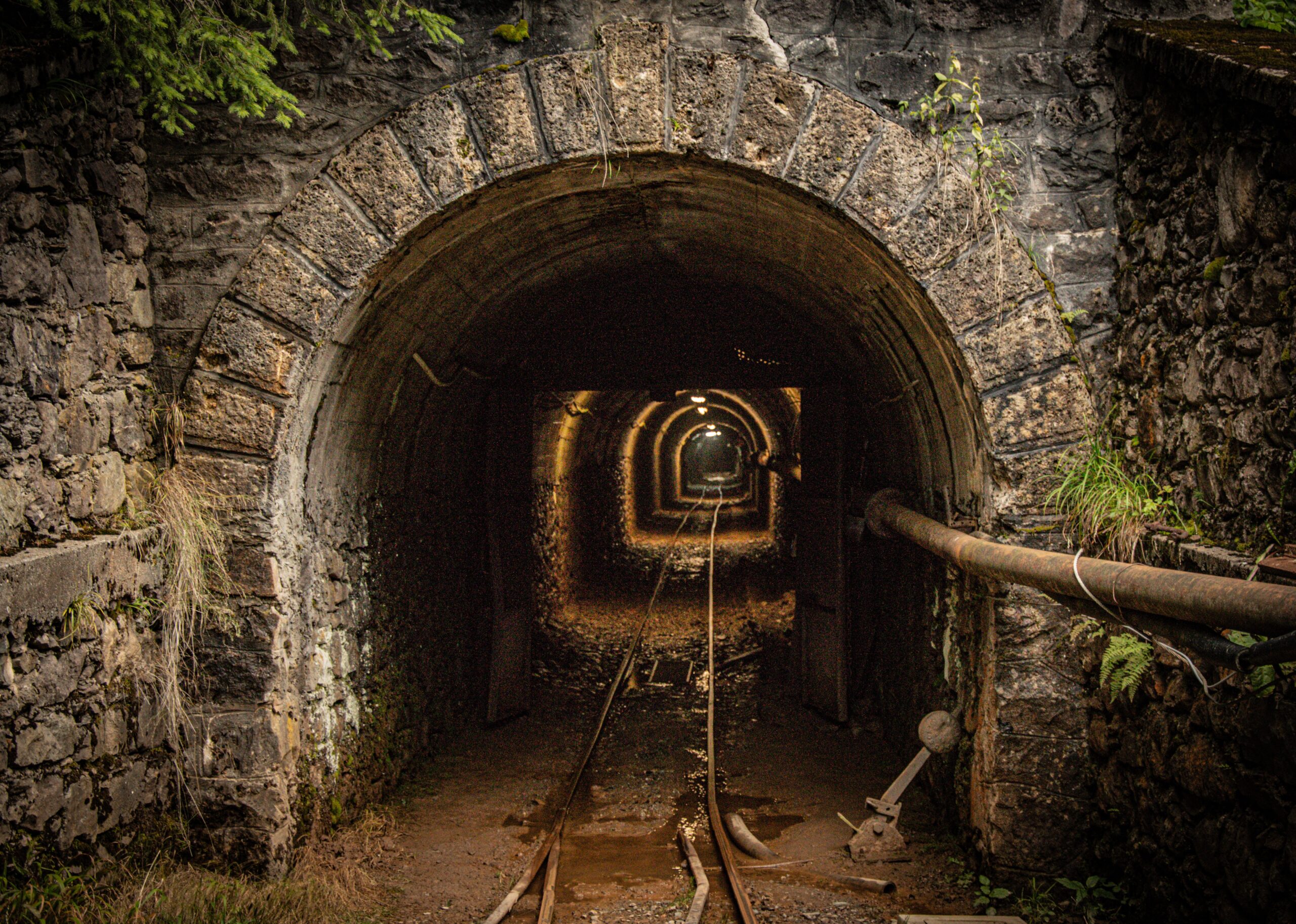 Entrance to a deep tunnel or mineshaft with some light rail tracks going deep into the vanishing point.
