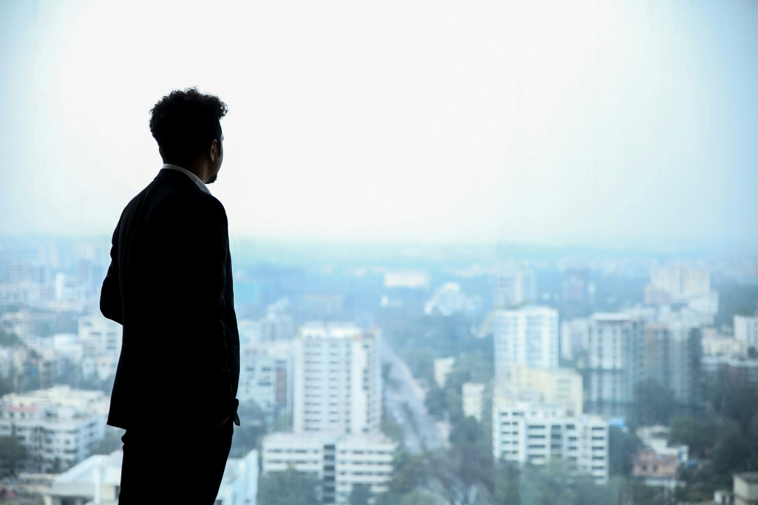 Person in silhouette wearing suit and looking out elevated window over a landscape with buildings and a cloudy sky.