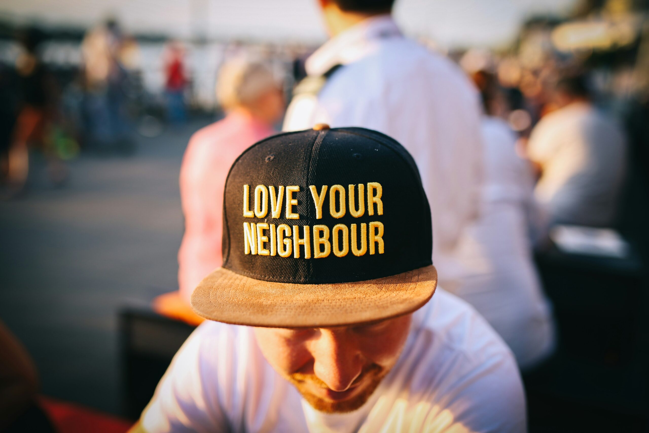 Man wearing a hat that says, "Love Your Neighbor" with some people blurry behind him.